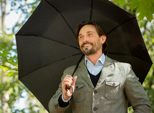 Un homme d'affaires en veste grise est sous un parapluie noir sus un temps ensoleillé. En arrière-plan, on peut apercevoir des arbres