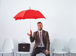 Un homme en costume et cravate assis sur une chaise tenant un parapluie rouge ouvert et sur une autre chaise est posé un attaché-case